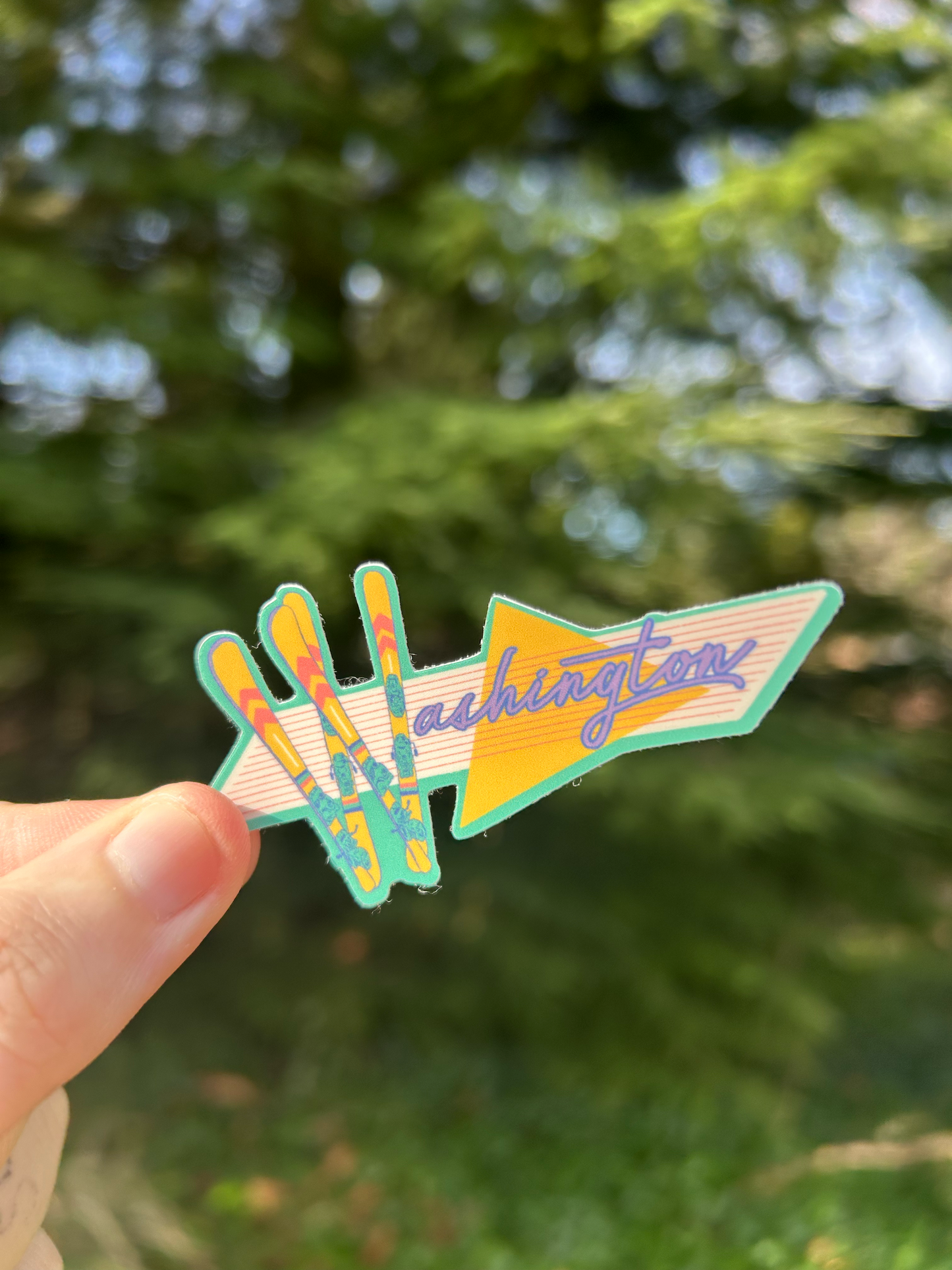Sticker with the word "Washington" on it, with the W letter made out of skis in a retro-vaporwave style being held up against a background of a pine tree.