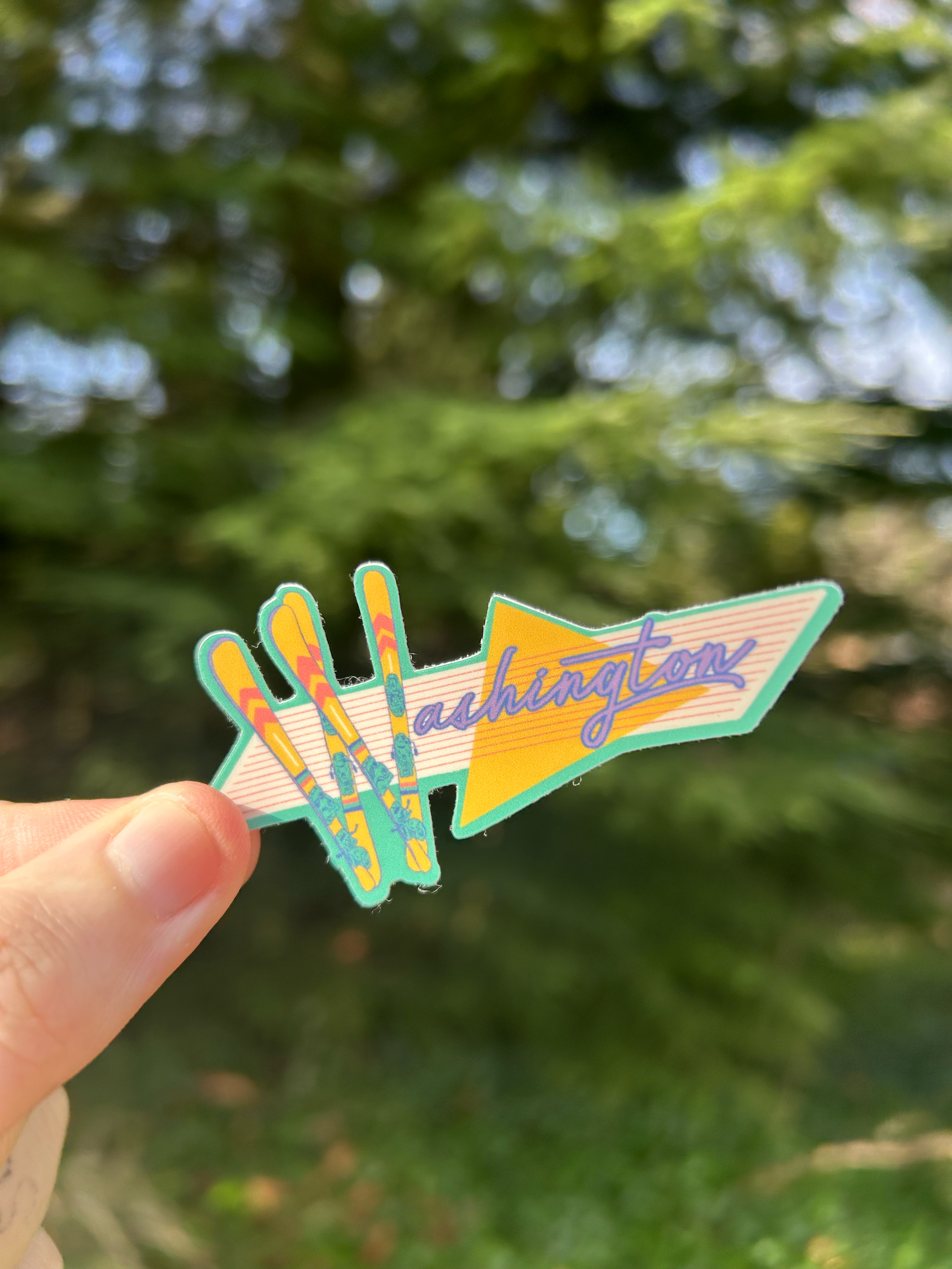 Sticker with the word "Washington" on it, with the W letter made out of skis in a retro-vaporwave style being held up against a background of a pine tree.